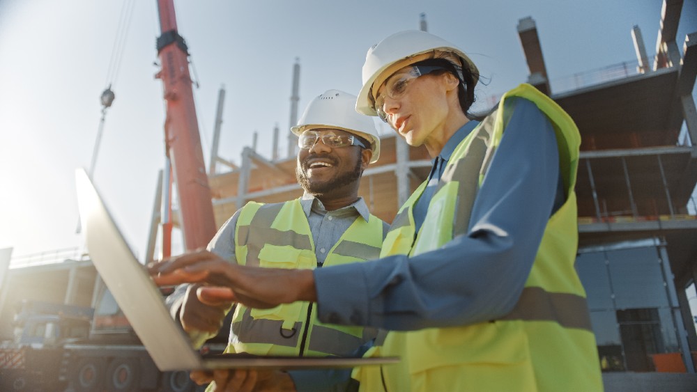 A male and female in hard hats and high vis jackets discussing plans on a laptop with a construction site in the background